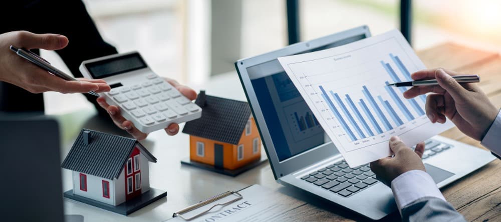 Two people discussing real estate data, with a calculator, model houses, and a bar chart on a laptop.