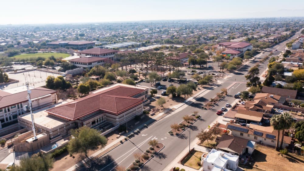 Aerial view of a suburban neighborhood with red-roofed buildings, roads, and scattered trees under a clear sky.