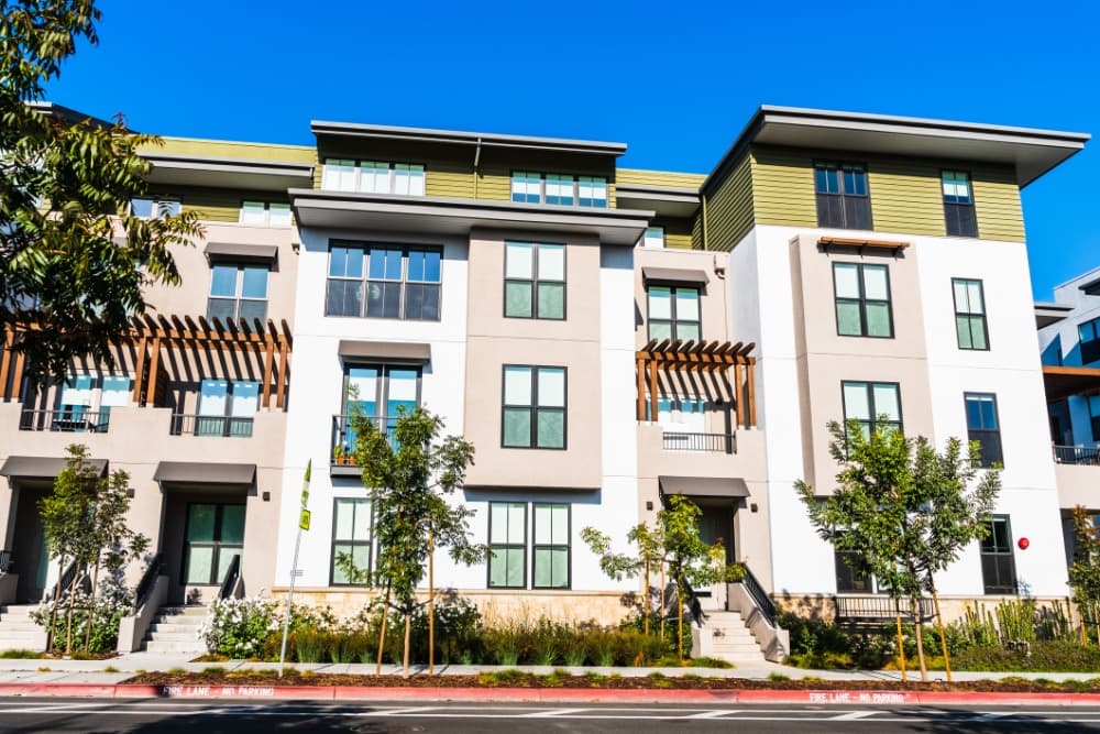 Modern multi-story apartment building with large windows, wooden accents, and surrounding trees under a clear blue sky.
