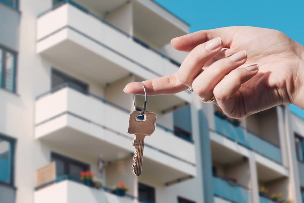A hand holding a key in front of a modern apartment building, symbolizing new home ownership or rental.