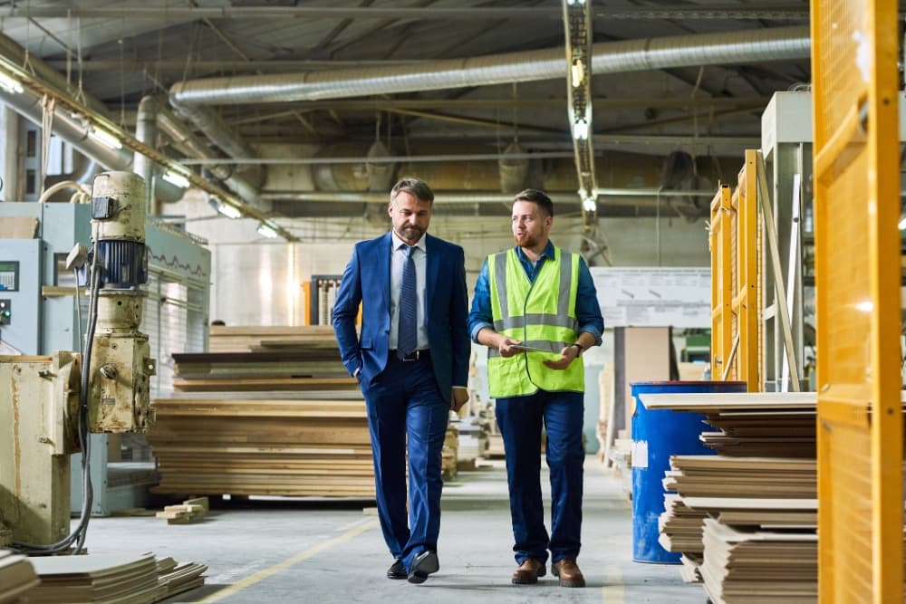 Two men walking in a factory; one in a suit, the other in a safety vest, surrounded by machinery and stacks of materials.