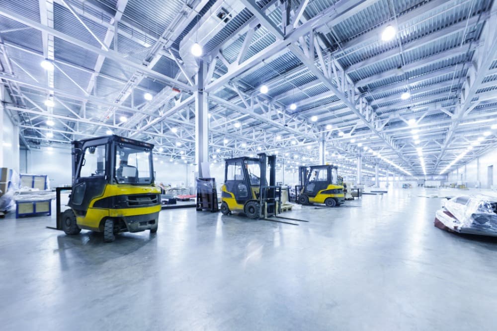 Spacious warehouse interior with high ceilings, bright lighting, and three yellow forklifts parked on the concrete floor.
