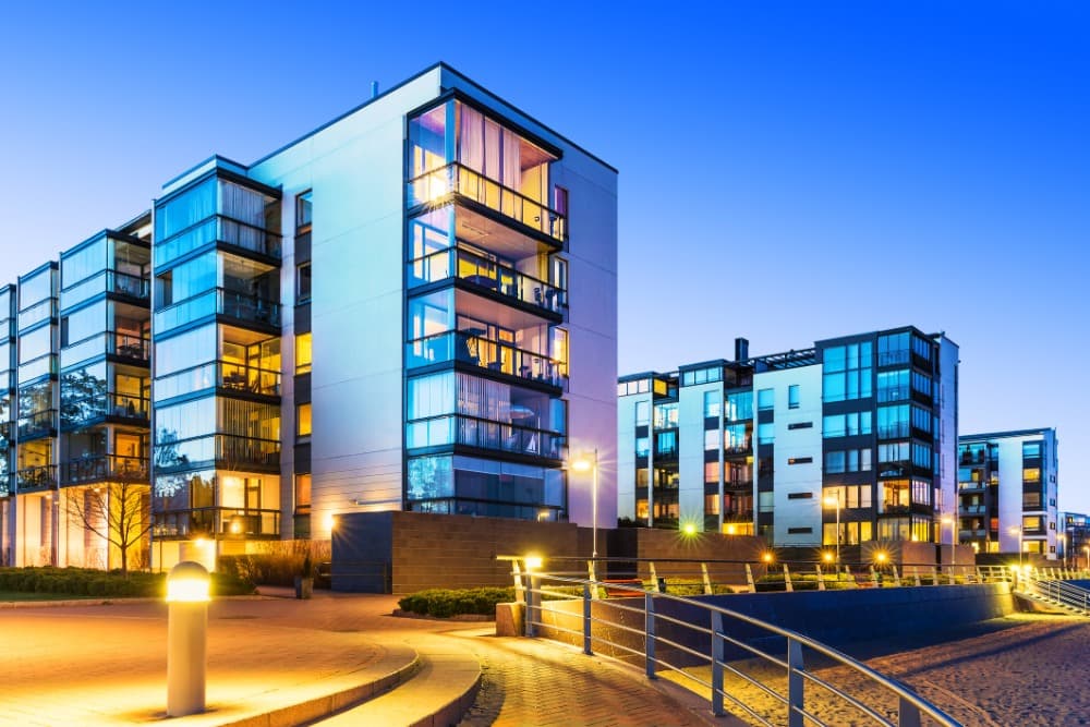 Modern apartment buildings with large windows and balconies, illuminated at dusk, surrounded by a paved walkway and contemporary railings.