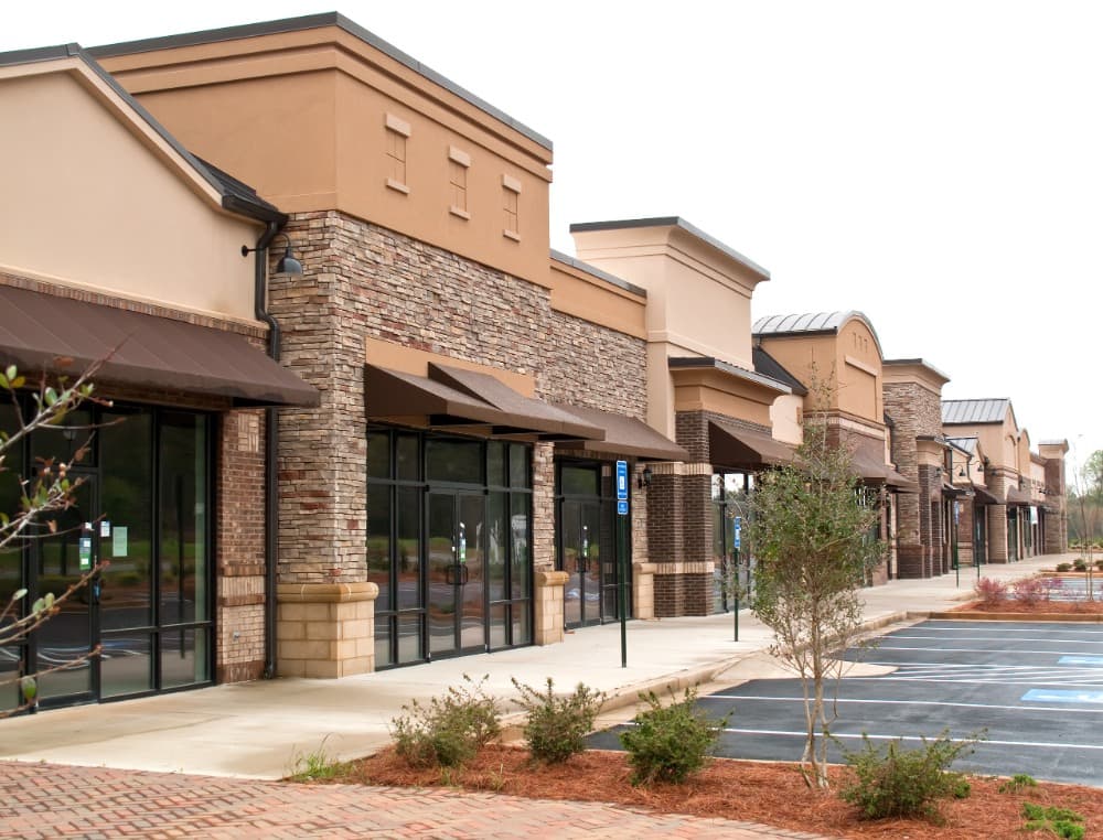 Empty strip mall with multiple storefronts, featuring brick and stone facades, large glass windows, and a mostly vacant parking lot.