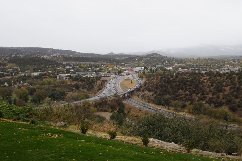 Overcast view of a highway curving through a hilly landscape, surrounded by greenery and scattered buildings in the distance.